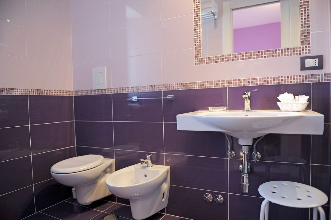 Modern bathroom with purple and pink tile design, wall-mounted sink, toilet, bidet, and illuminated mirror.