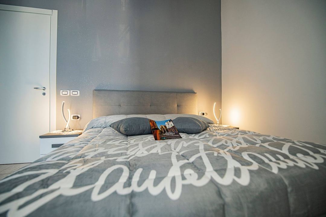 A modern bedroom featuring a gray upholstered headboard, patterned bedding, dark pillows, ambient lighting, and a minimalist design aesthetic.