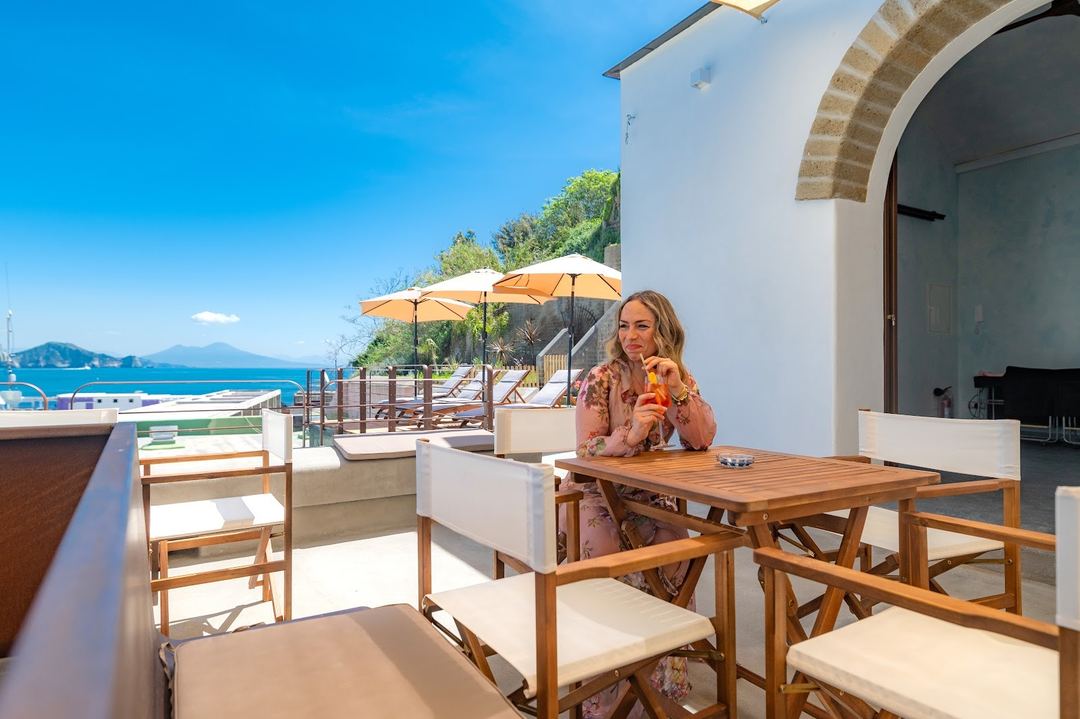 A guest enjoys refreshments on a seaside terrace overlooking the Mediterranean Sea with umbrellas and loungers visible in the background.