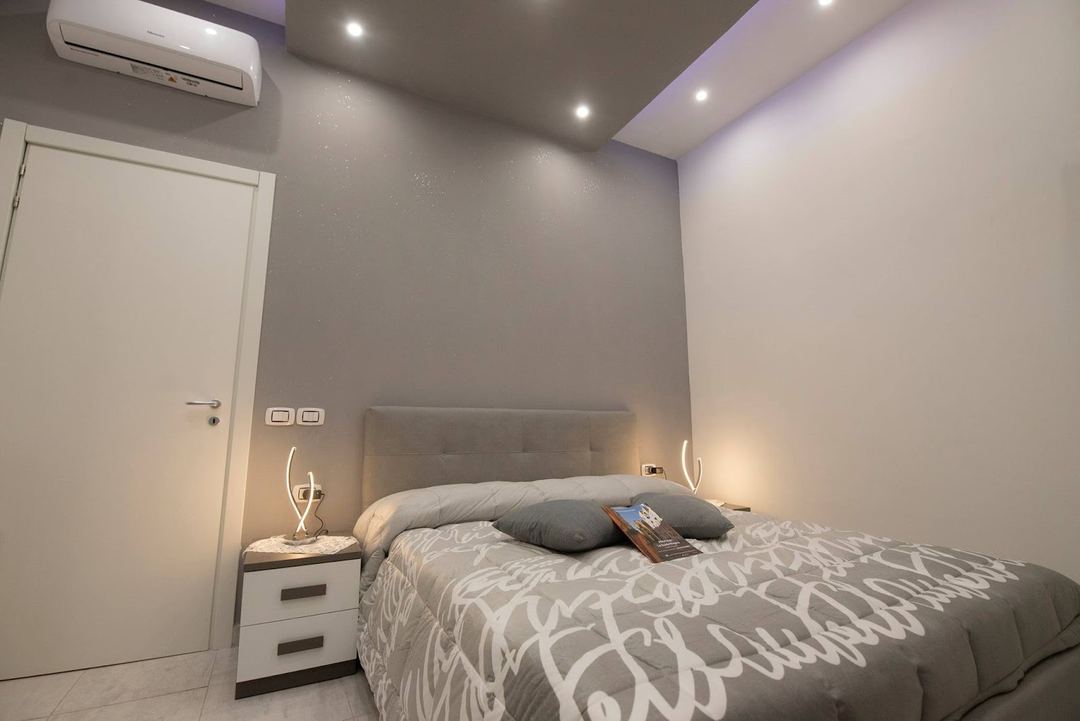 A modern bedroom featuring a gray upholstered headboard, patterned bedding, bedside tables with curved lighting fixtures, and ambient ceiling lights.
