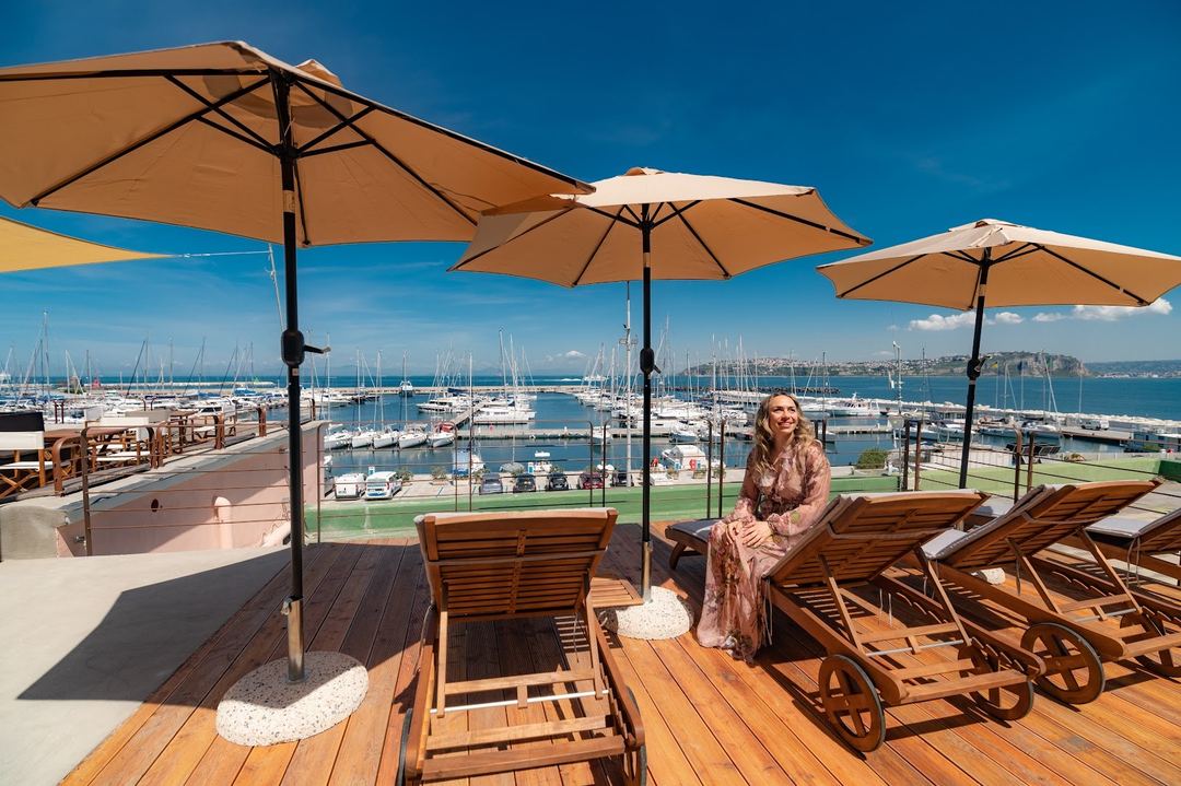 A woman relaxes on a wooden lounge chair under a tan umbrella on a terrace overlooking a marina filled with sailboats.