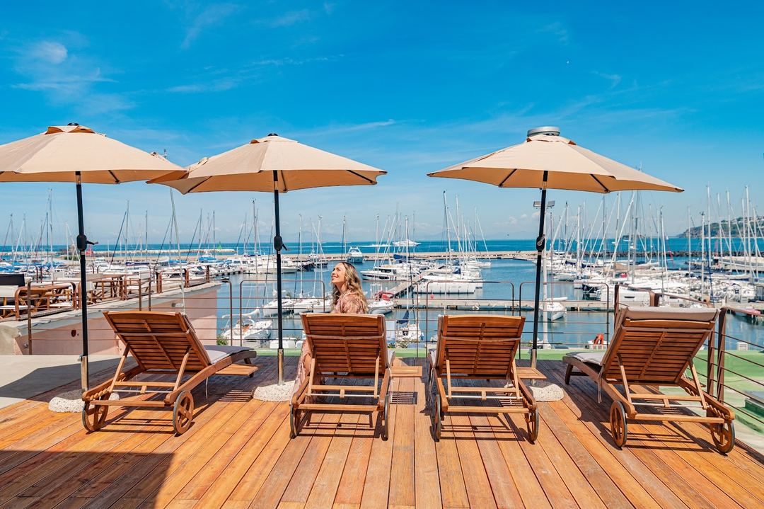 A waterfront terrace with wooden lounge chairs and umbrellas overlooking a marina filled with sailboats under a clear blue sky.