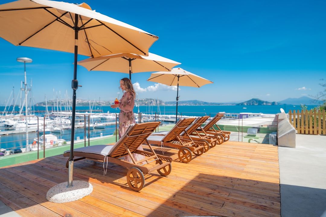 A woman relaxes on a wooden deck with sun loungers and umbrellas overlooking a marina and blue Mediterranean sea.