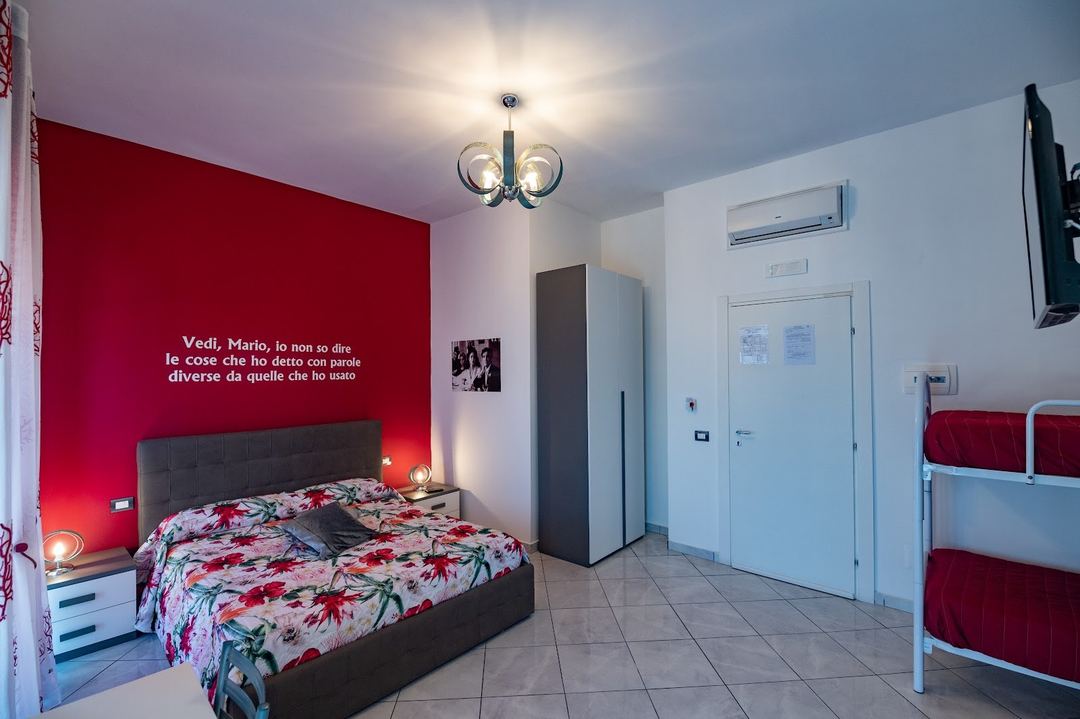 A modern bedroom with a bold red accent wall, black bed frame with floral bedding, and bunk beds visible on the right side.